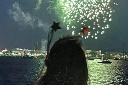 Person watching fireworks over water at night while holding a wand.